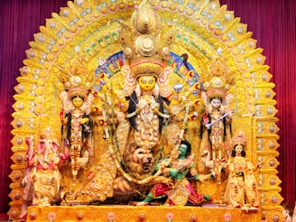 A richly decorated and colorful Durga Puja pandal displaying the goddess Durga with multiple arms, riding a lion, surrounded by other deities and figures adorned in vibrant garments and ornaments. The backdrop is intricately designed with golden and multicolored embellishments, creating a festive and majestic atmosphere.
