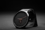 A sleek black wristwatch with a minimalist design sits against a dark, textured background. The watch features a black face with simple, thin hour markers and two delicate hands in a contrasting metallic color. A smaller subdial is positioned at the six o'clock mark. The watch strap is made of a black mesh material, adding to the overall elegant and modern aesthetic.