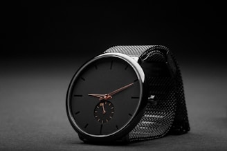 A sleek black wristwatch with a minimalist design sits against a dark, textured background. The watch features a black face with simple, thin hour markers and two delicate hands in a contrasting metallic color. A smaller subdial is positioned at the six o'clock mark. The watch strap is made of a black mesh material, adding to the overall elegant and modern aesthetic.
