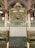 An ornate archway featuring intricate Arabic calligraphy in gold on a green background is seen, supported by pink and gold columns. There are decorative chandeliers and detailed patterns on the ceiling and walls. The floor is covered with a patterned green carpet.