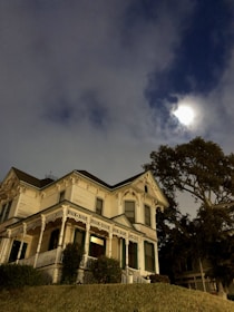 A large, ornate Victorian-style house with intricate architectural details is seen at night. The sky is partially cloudy with the moon glowing through, lending an eerie atmosphere. A large tree stands next to the house, and the house is set on a small hill with bushes and grass.