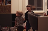 Two young children are playfully interacting in a cozy indoor setting. The room has a comfortable atmosphere with shelves of books and toys. A large armchair and some storage boxes are visible. Both children display expressions of joy and laughter.
