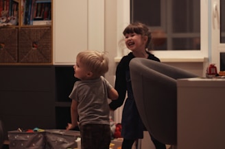 Two young children are playfully interacting in a cozy indoor setting. The room has a comfortable atmosphere with shelves of books and toys. A large armchair and some storage boxes are visible. Both children display expressions of joy and laughter.