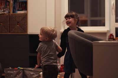 Two young children are playfully interacting in a cozy indoor setting. The room has a comfortable atmosphere with shelves of books and toys. A large armchair and some storage boxes are visible. Both children display expressions of joy and laughter.