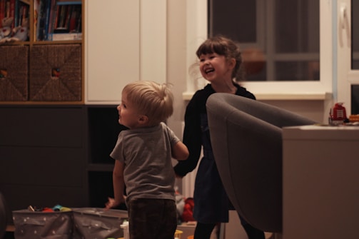 Two young children are playfully interacting in a cozy indoor setting. The room has a comfortable atmosphere with shelves of books and toys. A large armchair and some storage boxes are visible. Both children display expressions of joy and laughter.