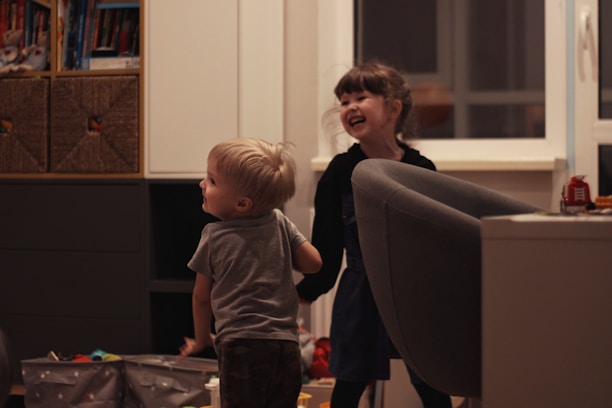 Two young children are playfully interacting in a cozy indoor setting. The room has a comfortable atmosphere with shelves of books and toys. A large armchair and some storage boxes are visible. Both children display expressions of joy and laughter.
