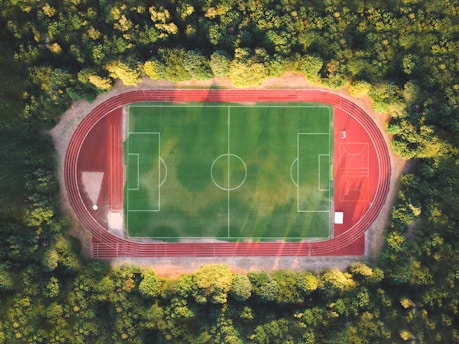 An aerial view of a soccer field with a surrounding red running track, nestled within a dense forest. The vibrant green of the field contrasts with the deep red of the track and the lush greenery of the surrounding trees.