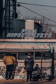 A small street vendor stall with a mural of underwater scenes is attended by two people standing in front. The sign above the stall has Cyrillic lettering, giving it a distinctly local feel. Weathered and industrial structures frame the background along with some leafless trees and suspended cables.
