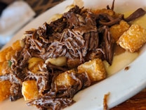 A close-up view of a dish featuring tender, shredded beef combined with crispy, golden-brown potato cubes on a white plate. The dish appears to have a creamy sauce beneath the beef and potatoes.