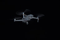 A drone with four rotors is captured in flight against a dark background. The drone's design is sleek and metallic, and it appears to be hovering steadily.