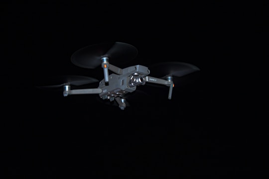 A drone with four rotors is captured in flight against a dark background. The drone's design is sleek and metallic, and it appears to be hovering steadily.
