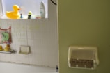 A bathroom scene with various rubber ducks placed on a windowsill and bathtub area. The white tiles have green text saying 'I feel fine' and some hand-drawn decorations above. A mesh bag hanging from the wall contains bath toys. An empty toilet paper roll in a holder has the handwritten text 'IT'S THE END OF THE ROLL AS WE KNOW IT!' on it.