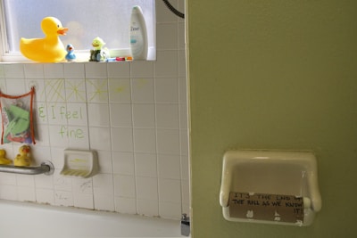 A bathroom scene with various rubber ducks placed on a windowsill and bathtub area. The white tiles have green text saying 'I feel fine' and some hand-drawn decorations above. A mesh bag hanging from the wall contains bath toys. An empty toilet paper roll in a holder has the handwritten text 'IT'S THE END OF THE ROLL AS WE KNOW IT!' on it.