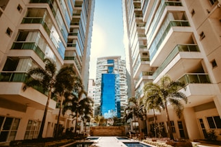 A modern urban setting with tall apartment buildings flanking a central courtyard area. The buildings have multiple floors with reflective glass windows and balconies. Palm trees are planted along the pathways, and there is a narrow swimming pool in the foreground. In the distance, a taller building with a distinct blue facade stands prominently.