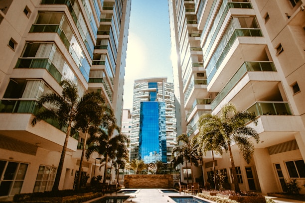 A modern urban setting with tall apartment buildings flanking a central courtyard area. The buildings have multiple floors with reflective glass windows and balconies. Palm trees are planted along the pathways, and there is a narrow swimming pool in the foreground. In the distance, a taller building with a distinct blue facade stands prominently.