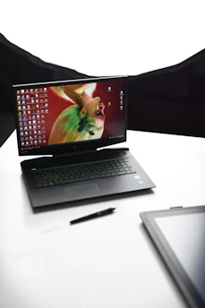 A laptop with an artistic wallpaper on the screen, featuring a colorful, abstract portrait. The laptop is angled and placed on a clean, white surface. A pen lies beside the laptop, and the corner of another black electronic device is visible in the foreground.
