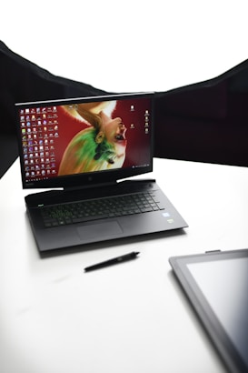 A laptop with an artistic wallpaper on the screen, featuring a colorful, abstract portrait. The laptop is angled and placed on a clean, white surface. A pen lies beside the laptop, and the corner of another black electronic device is visible in the foreground.