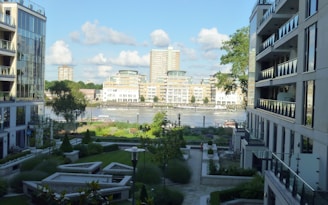A modern urban scene with high-rise buildings flanking a central courtyard. The landscape features well-manicured greenery and pathways, with a river visible in the background. The architecture is sleek and contemporary, with large windows reflecting the bright daylight. The sky is a clear blue with some fluffy clouds.