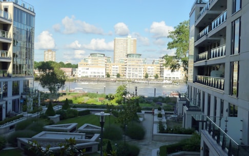 A modern urban scene with high-rise buildings flanking a central courtyard. The landscape features well-manicured greenery and pathways, with a river visible in the background. The architecture is sleek and contemporary, with large windows reflecting the bright daylight. The sky is a clear blue with some fluffy clouds.