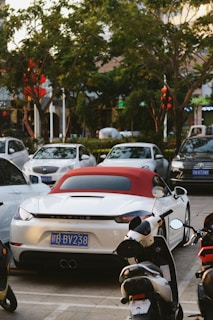 A white Porsche sports car with a red convertible top is parked in a lot alongside other cars and motorbikes. The background includes trees and a building with some visible shop signs.