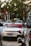 A white Porsche sports car with a red convertible top is parked in a lot alongside other cars and motorbikes. The background includes trees and a building with some visible shop signs.