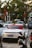 A white Porsche sports car with a red convertible top is parked in a lot alongside other cars and motorbikes. The background includes trees and a building with some visible shop signs.