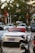 A white Porsche sports car with a red convertible top is parked in a lot alongside other cars and motorbikes. The background includes trees and a building with some visible shop signs.