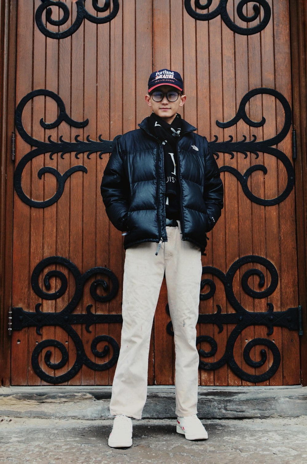 man in black leather jacket and white pants standing beside brown wooden wall