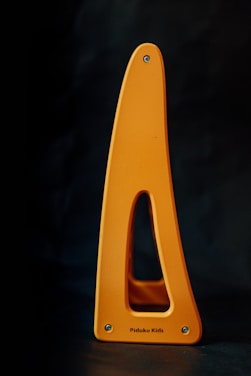 A yellow-orange triangular object with a cutout in the center, featuring small screws at its base and a smooth surface. The words 'Pidoko Kids' are printed near the bottom edge.