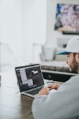 A person wearing a gray hoodie and a white cap is sitting at a wooden table, working on a laptop. The screen displays a video editing software with various windows open. The environment appears to be a casual, home setting with a blurred television in the background.