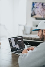 A person wearing a gray hoodie and a white cap is sitting at a wooden table, working on a laptop. The screen displays a video editing software with various windows open. The environment appears to be a casual, home setting with a blurred television in the background.