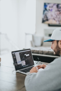 A person wearing a gray hoodie and a white cap is sitting at a wooden table, working on a laptop. The screen displays a video editing software with various windows open. The environment appears to be a casual, home setting with a blurred television in the background.