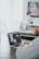 A person wearing a gray hoodie and a white cap is sitting at a wooden table, working on a laptop. The screen displays a video editing software with various windows open. The environment appears to be a casual, home setting with a blurred television in the background.