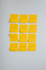 Twelve yellow sticky notes are arranged in a grid on a light gray wall. Each note contains a part of a message written in black ink, which together form a religious or inspirational quote.