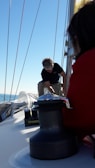 A person is working on a sailboat, adjusting or handling equipment on the deck. The mast extends up with lines running parallel to it. The backdrop includes a clear blue sky and a glimpse of ocean. Another person is partially visible in the foreground, wearing a red garment.