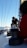 A person is working on a sailboat, adjusting or handling equipment on the deck. The mast extends up with lines running parallel to it. The backdrop includes a clear blue sky and a glimpse of ocean. Another person is partially visible in the foreground, wearing a red garment.