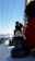 A person is working on a sailboat, adjusting or handling equipment on the deck. The mast extends up with lines running parallel to it. The backdrop includes a clear blue sky and a glimpse of ocean. Another person is partially visible in the foreground, wearing a red garment.