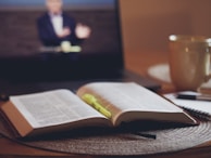 An open book with marked pages lies on a table, accompanied by a yellow highlighter pen. In the background, a laptop screen displays a blurred image of a person, suggesting a video call or online lecture. A large mug and a notebook with a pen are also visible on the table, creating an atmosphere of study or work from home.