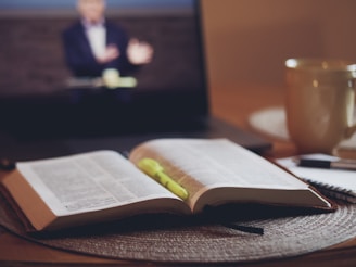 An open book with marked pages lies on a table, accompanied by a yellow highlighter pen. In the background, a laptop screen displays a blurred image of a person, suggesting a video call or online lecture. A large mug and a notebook with a pen are also visible on the table, creating an atmosphere of study or work from home.