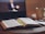 An open book with marked pages lies on a table, accompanied by a yellow highlighter pen. In the background, a laptop screen displays a blurred image of a person, suggesting a video call or online lecture. A large mug and a notebook with a pen are also visible on the table, creating an atmosphere of study or work from home.