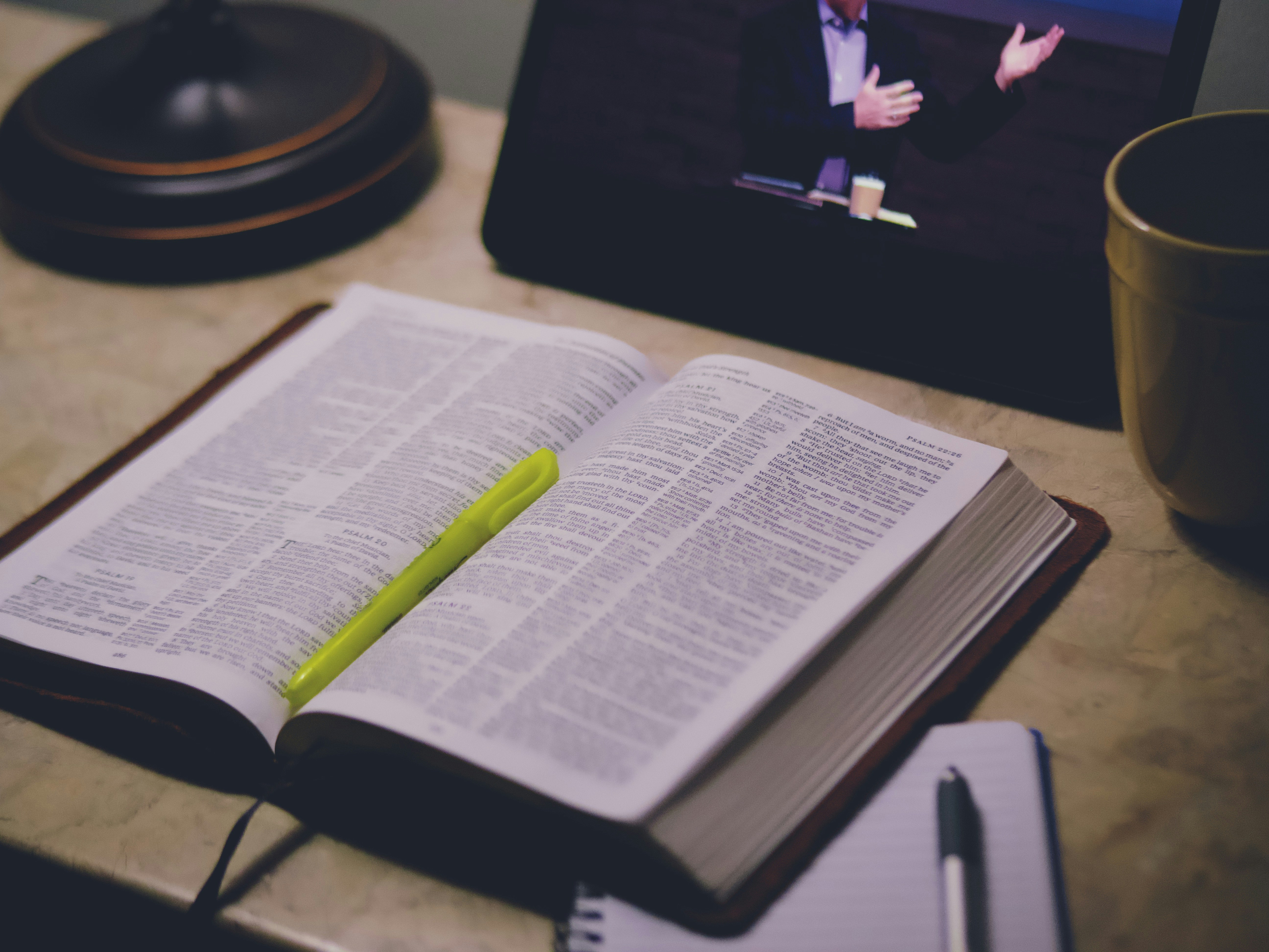 An open Bible with a yellow highlighter in the foreground with an ipad with a preacher in the background.