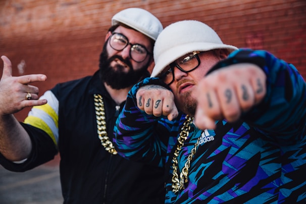Two individuals appear in streetwear fashion with bold accessories. They are wearing distinctive hats and sporting large gold chains. Tattoos with numbers are visible on the fist of one individual, while the other has a ring. Both have eyeglasses, and their attire includes bold patterns and logos.