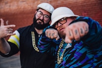 Two individuals appear in streetwear fashion with bold accessories. They are wearing distinctive hats and sporting large gold chains. Tattoos with numbers are visible on the fist of one individual, while the other has a ring. Both have eyeglasses, and their attire includes bold patterns and logos.
