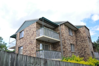 A multi-story residential building with a brick facade and white metal railings on the balconies. The building is surrounded by greenery, such as shrubs and trees, and there's a wooden fence in the foreground. The sky is partly cloudy, creating a bright and airy atmosphere.