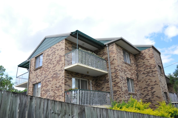 A multi-story residential building with a brick facade and white metal railings on the balconies. The building is surrounded by greenery, such as shrubs and trees, and there's a wooden fence in the foreground. The sky is partly cloudy, creating a bright and airy atmosphere.