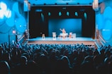 A theater setting with a performer on stage in front of a packed audience. The stage is lit with blue lighting, highlighting the central performer who is standing and engaging with the audience. There are props on stage, including chairs and a table.