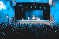 A theater setting with a performer on stage in front of a packed audience. The stage is lit with blue lighting, highlighting the central performer who is standing and engaging with the audience. There are props on stage, including chairs and a table.