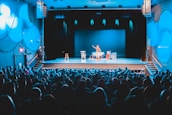 A theater setting with a performer on stage in front of a packed audience. The stage is lit with blue lighting, highlighting the central performer who is standing and engaging with the audience. There are props on stage, including chairs and a table.