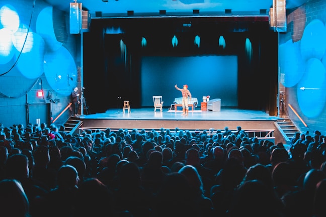 A theater setting with a performer on stage in front of a packed audience. The stage is lit with blue lighting, highlighting the central performer who is standing and engaging with the audience. There are props on stage, including chairs and a table.