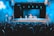 A theater setting with a performer on stage in front of a packed audience. The stage is lit with blue lighting, highlighting the central performer who is standing and engaging with the audience. There are props on stage, including chairs and a table.